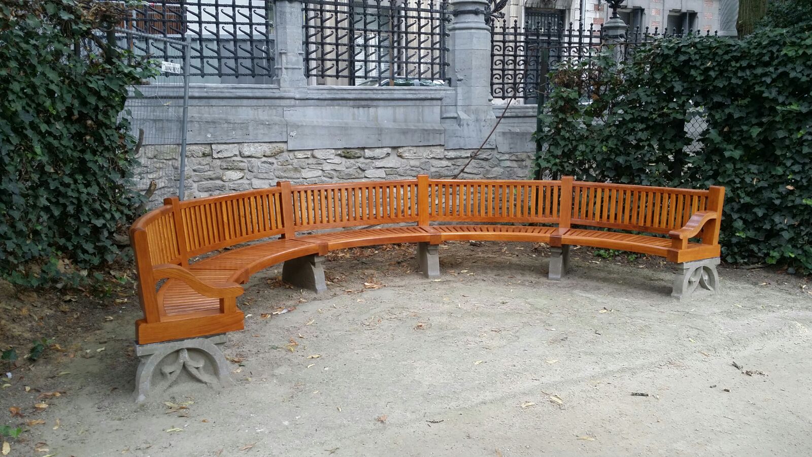 Un nouveau banc en bois semi-circulaire placé devant l'un des anciens murs de clôture de la place du Petit Sablon. Le nouveau banc a un style classique, identique aux bancs originaux du parc.