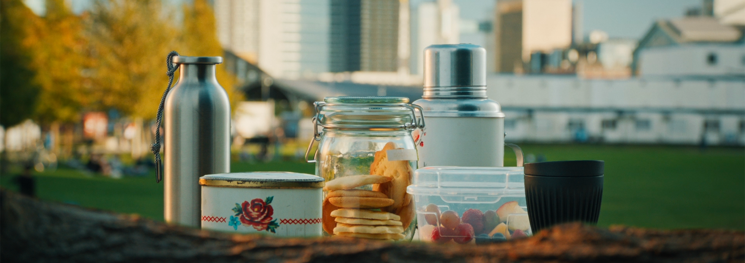 Une gourde et un thermos en acier inoxydable, un bocal en verre, une boîte à biscuits en métal, un tupperware en plastique et une tasse réutilisable sur une table dans un parc.