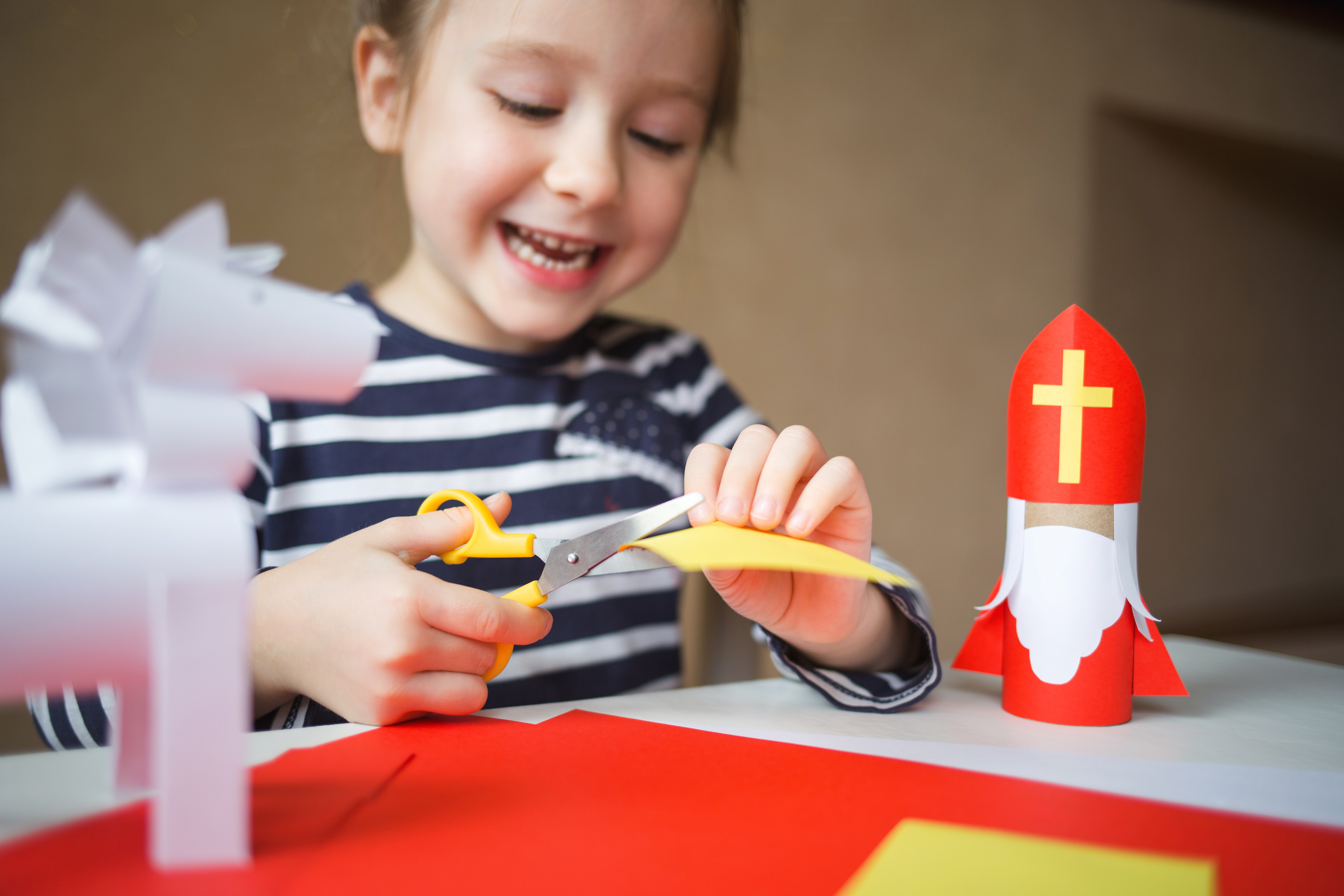 Une fille fabrique Saint-Nicolas avec un rouleau de papier toilette vide et du papier coloré. Elle découpe un morceau de papier jaune avec des ciseaux.