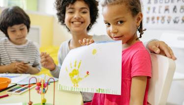 Une petite fille montre un dessin dans une classe, son institutrice sourit en arrière-plan